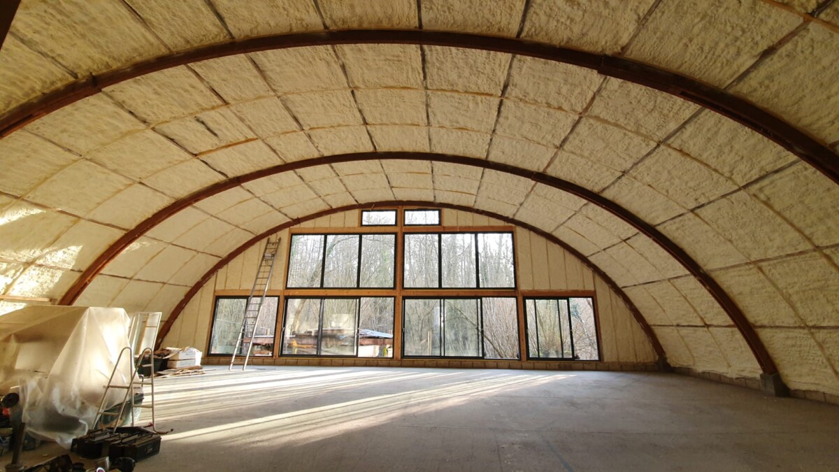 isolatie-wanden-interieurs Binnenin een groot gebouw met een gewelfde houten dakstructuur, geïsoleerd met spray-schuim. Een groot panoramisch raam biedt uitzicht op de kale bomen buiten.