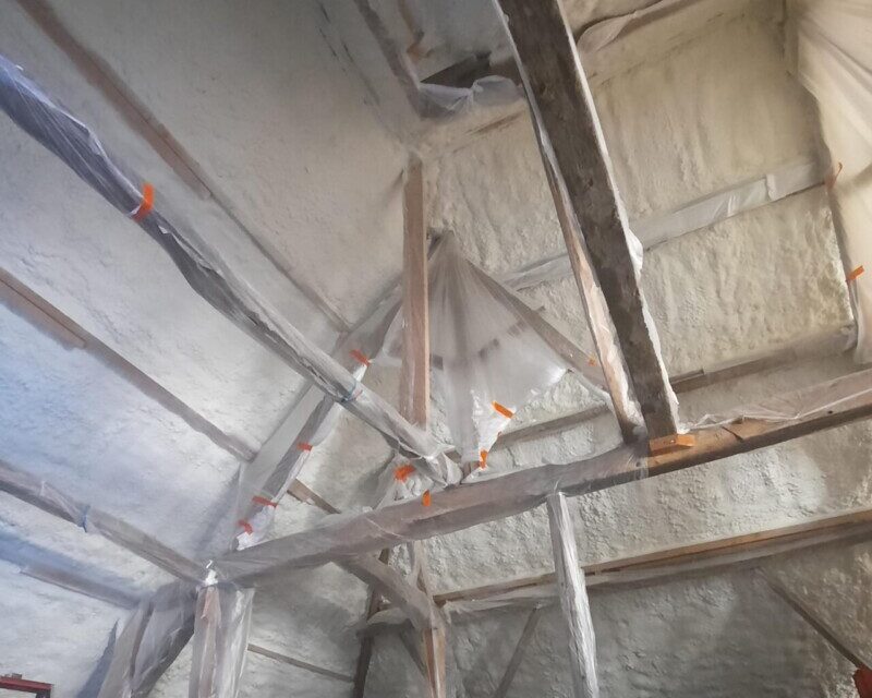 Isolation de la toiture intérieure d'une grange à Affligem, avec des poutres apparentes et de la mousse isolante projetée.