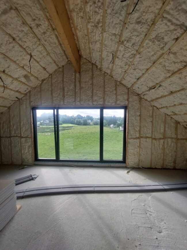 Grande pièce avec plafond en pente, isolée par de la mousse projetée entre les chevrons en bois visibles. Une grande baie vitrée coulissante donne sur un vaste paysage de prairies.