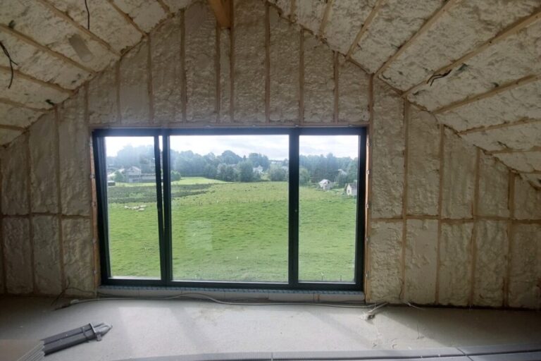 Grande pièce avec plafond en pente, isolée par de la mousse projetée entre les chevrons en bois visibles. Une grande baie vitrée coulissante donne sur un vaste paysage de prairies.