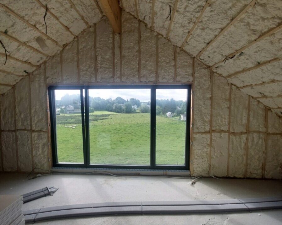 Grande pièce avec plafond en pente, isolée par de la mousse projetée entre les chevrons en bois visibles. Une grande baie vitrée coulissante donne sur un vaste paysage de prairies.