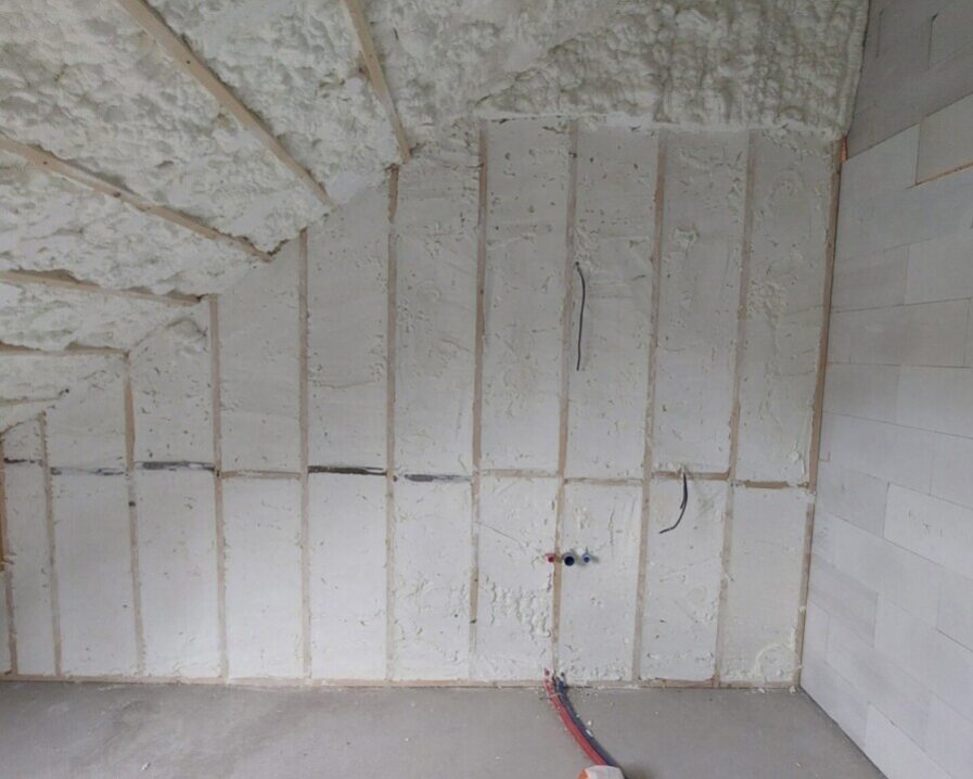 Mur incliné d'une pièce, isolé avec de la mousse projetée. Des gaines électriques et de plomberie sont visibles au sol.