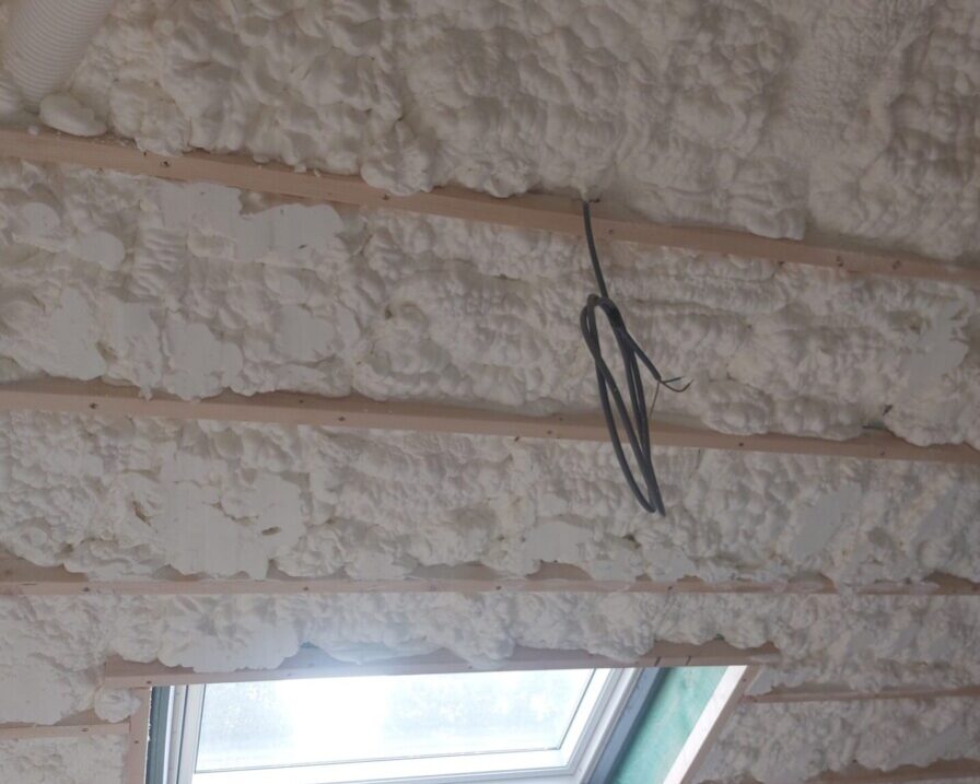 Plafond en pente isolé avec mousse polyuréthane projetée entre des chevrons en bois, avec plusieurs fils électriques apparents, dans une pièce rénovée de ferme.