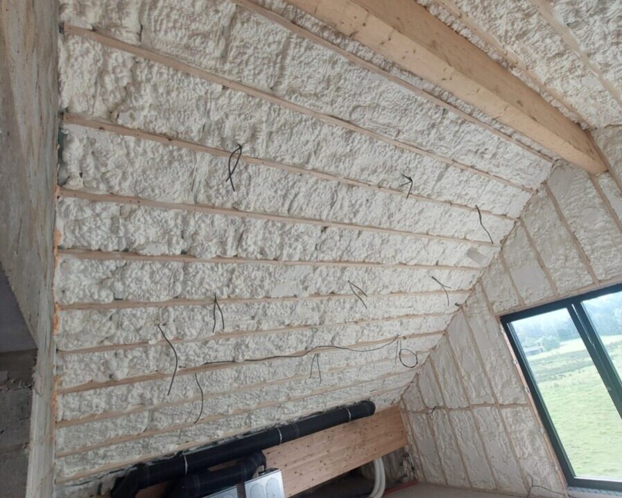Plafond en pente isolé avec de la mousse projetée, chevrons en bois visibles, câblage électrique apparent, et système de ventilation installé dans une pièce en rénovation.