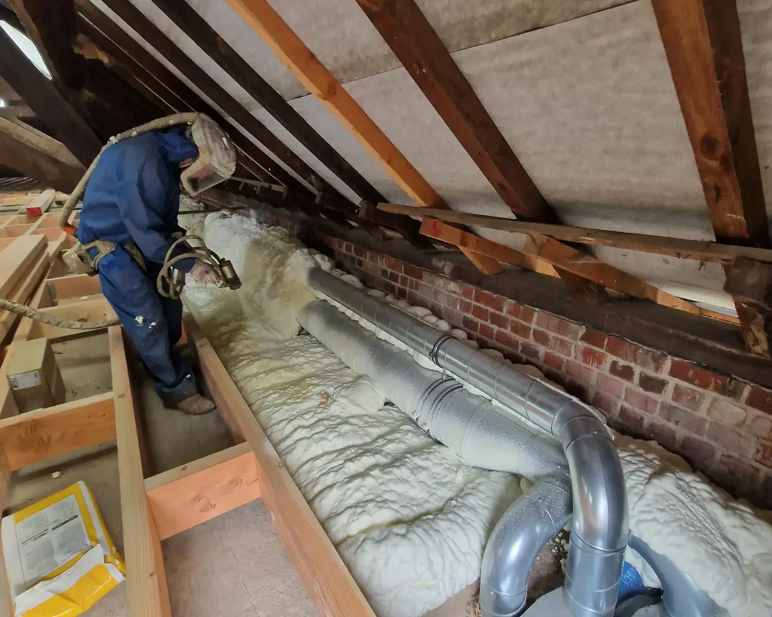 Application de mousse ICYNENE LITE+ autour des tuyaux de ventilation et de chauffage dans le grenier d'une maison à Destelbergen, garantissant une isolation thermique et acoustique optimale.