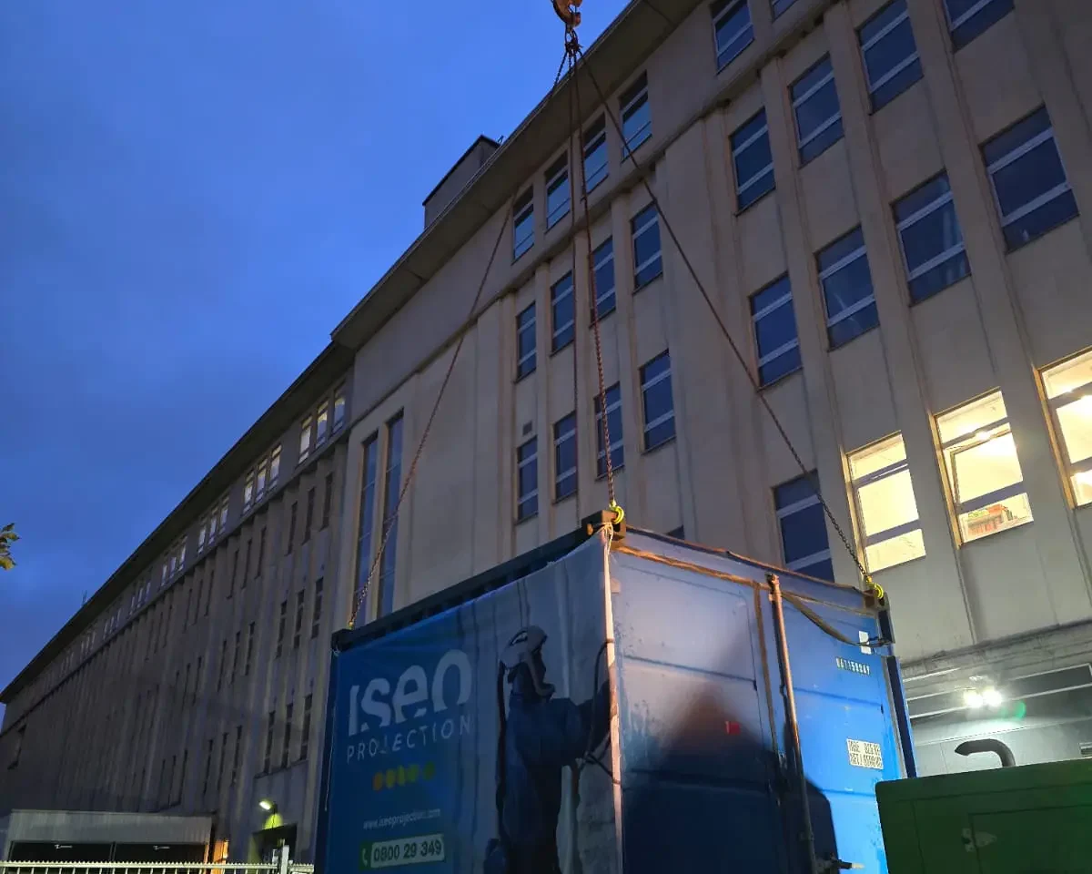 Grue déposant un conteneur ISEO Projection dans la cour intérieure d’un immeuble à Forest, solution logistique adaptée pour isoler une toiture plate et des coursives malgré les contraintes d'accès.