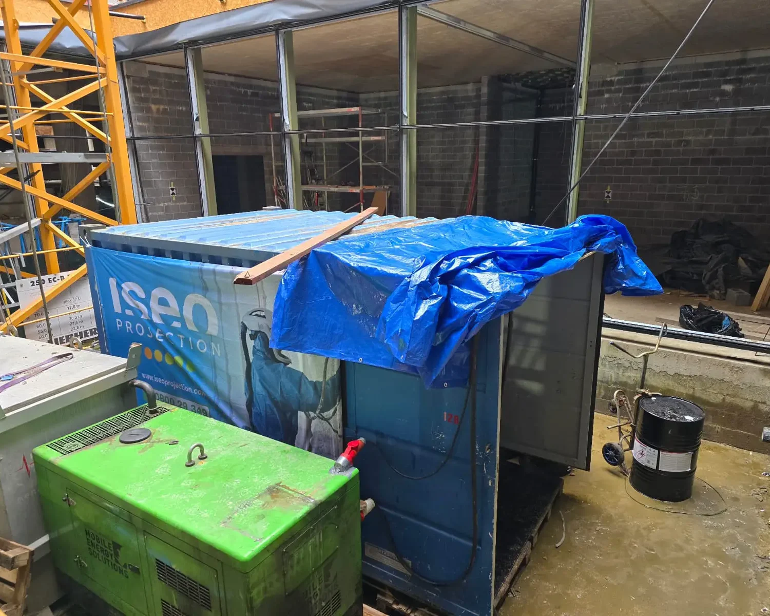 Conteneur bleu avec l’unité de projection ISEO sécurisé par une bâche bleue sur le chantier d’isolation à Forest, entouré d’équipements techniques.