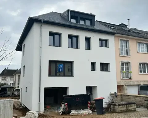 Maison à trois étages récemment isolée à Howald, Luxembourg, avec une façade blanche et une toiture rénovée. Les finitions de chantier sont en cours, avec du matériel et des barrières visibles à l’avant du bâtiment.