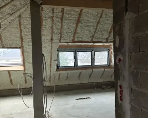Combles en cours de rénovation avec isolation en mousse projetée sur la toiture intérieure. L’espace est encore en chantier, avec des fils électriques apparents et des ouvertures vitrées apportant de la lumière naturelle.