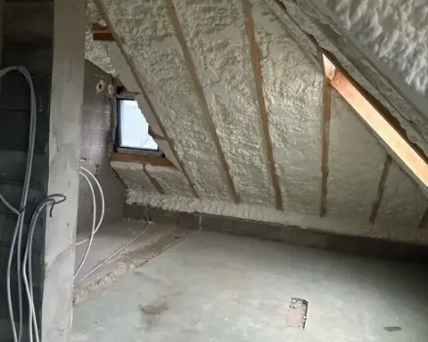 Isolation de toiture en mousse projetée appliquée sur la sous-pente d’un comble en rénovation. L’espace montre la structure apparente avec des poutres et des fils électriques en attente d’installation.