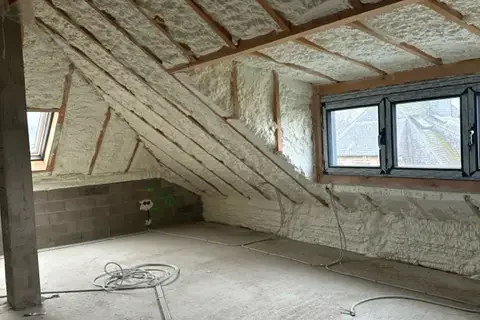 Combles aménageables après application d'une isolation en mousse projetée sur la toiture. L’espace lumineux comprend plusieurs fenêtres et une structure en bois visible sous l’isolant.