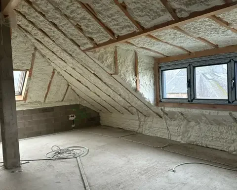 Combles aménageables après application d'une isolation en mousse projetée sur la toiture. L’espace lumineux comprend plusieurs fenêtres et une structure en bois visible sous l’isolant.
