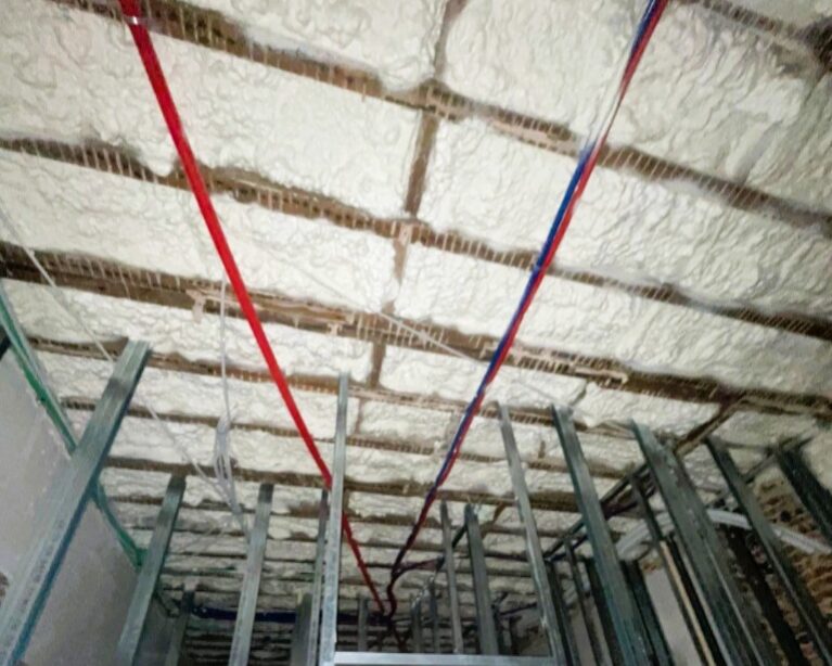 Isolation de plafond en mousse polyuréthane H2O projetée (12 cm) entre solives d'une maison en rénovation à Zaventem, avec gaines techniques et ossature métallique apparentes.