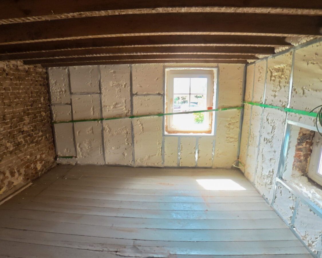 Pièce rénovée d’une fermette de 1850 avec murs irréguliers isolés par mousse polyuréthane projetée entre une structure métallique et des poutres apparentes.